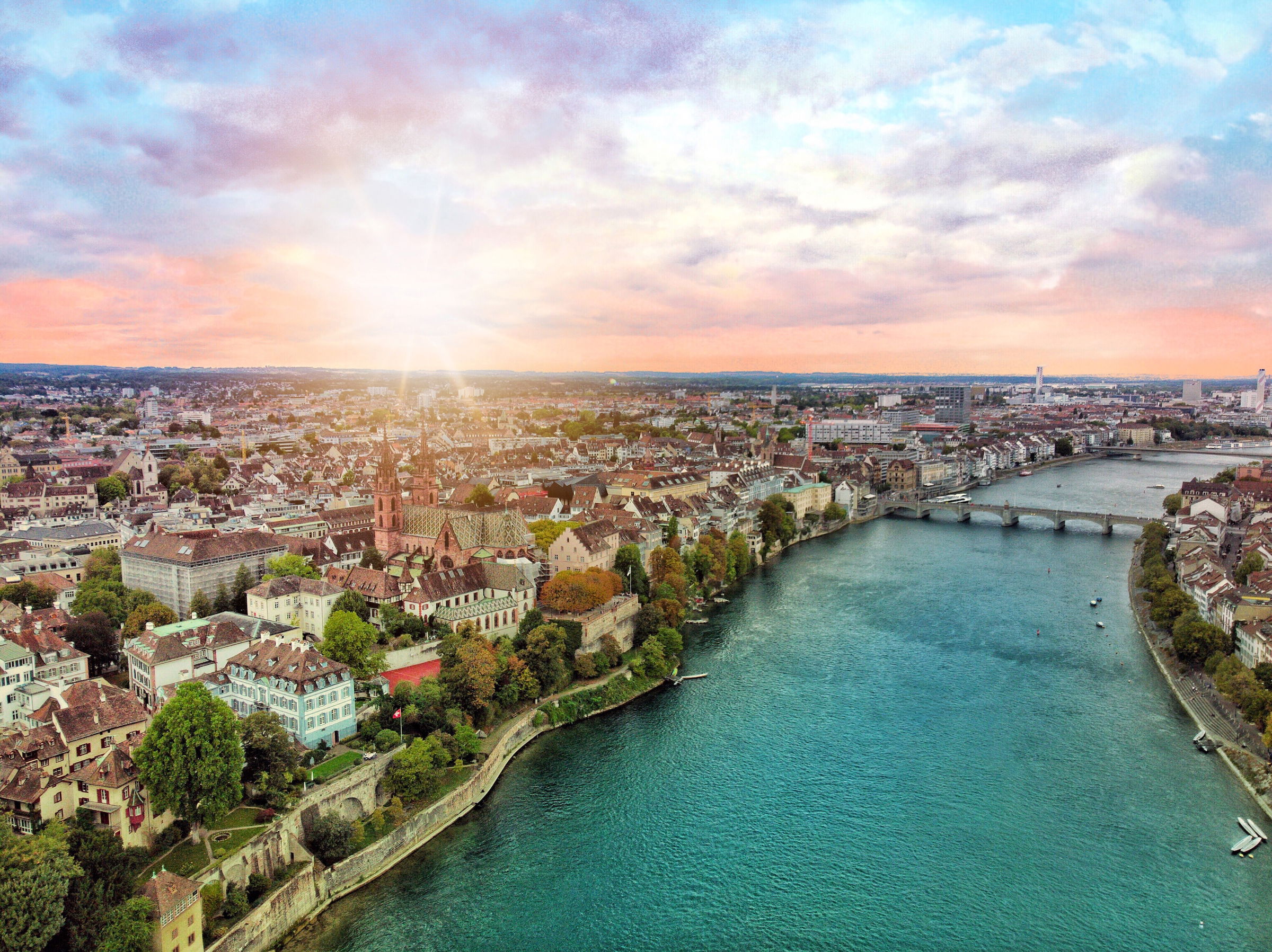 Luftaufnahme der Stadt Basel bei Sonnenuntergang mit dem Rhein in der Bildmitte und historischen Gebäuden am Ufer.