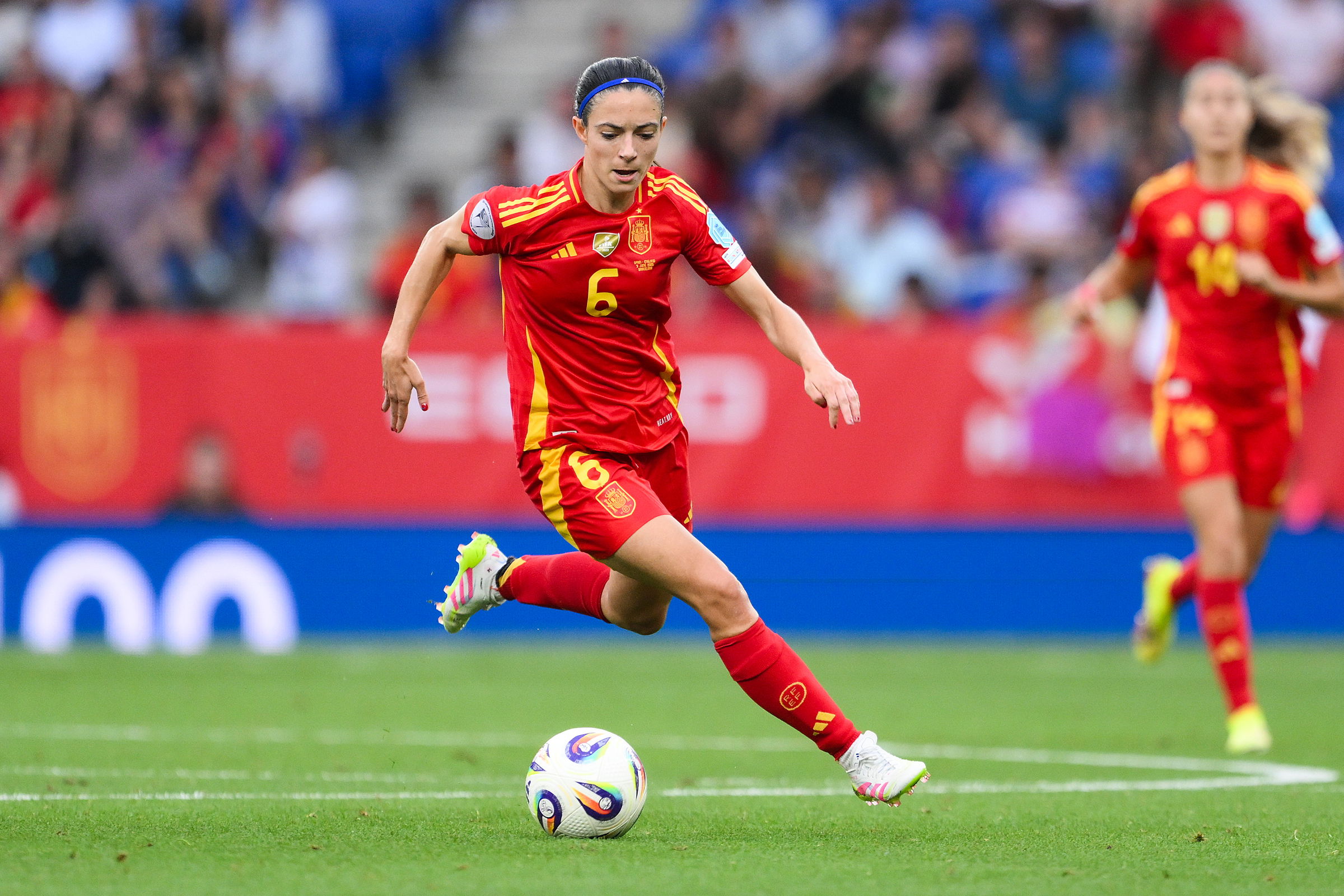Spanische Fußballspielerin in rotem Trikot mit der Nummer 6 dribbelt den Ball während eines Spiels auf dem Spielfeld. Im Hintergrund sind Mitspielerin und Zuschauer zu sehen.