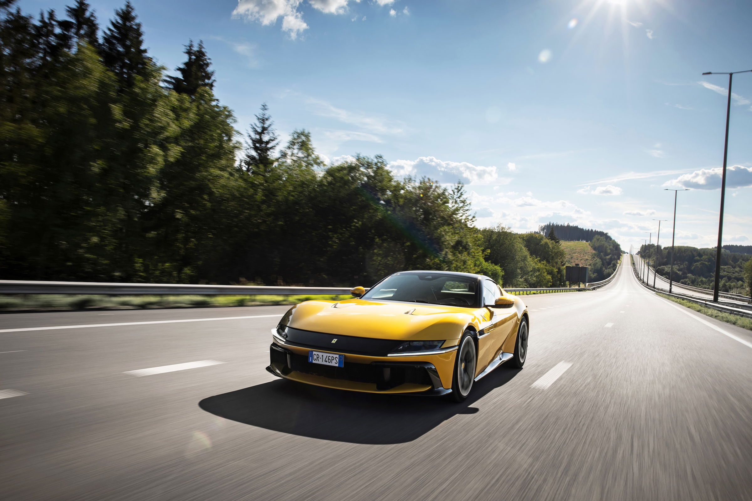 Gelber Ferrari 12Cilindri fährt auf leerer Landstraße bei Sonnenschein, umgeben von Bäumen und leicht hügeliger Landschaft.
