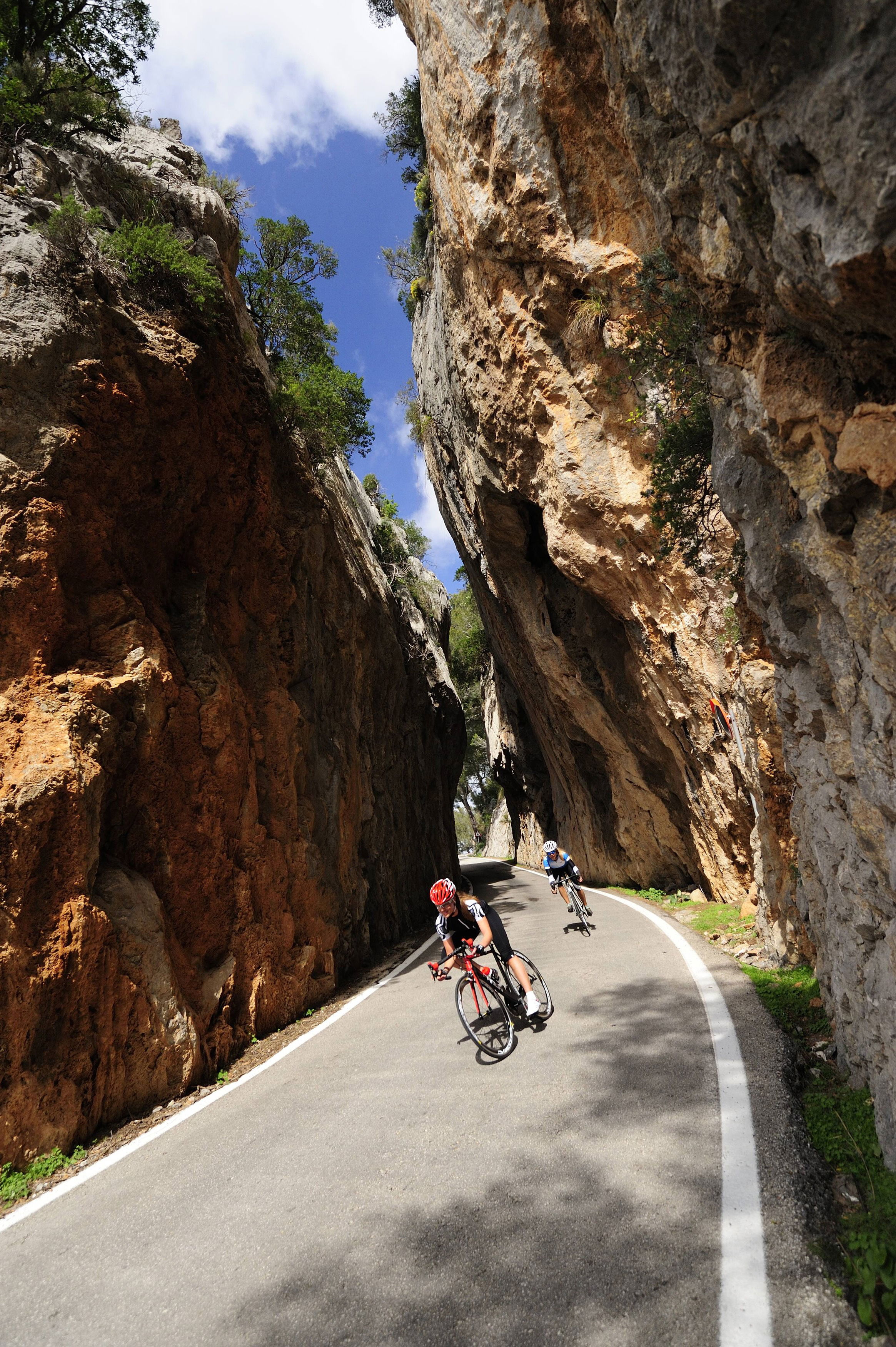 Zwei Rennradfahrer durchqueren eine enge Felsenschlucht auf Mallorca. Die Straße schlängelt sich zwischen steil aufragenden Kalksteinfelsen hindurch, darüber blauer Himmel mit einigen Wolken. Die Szene vermittelt sportliche Dynamik und beeindruckende Natur.