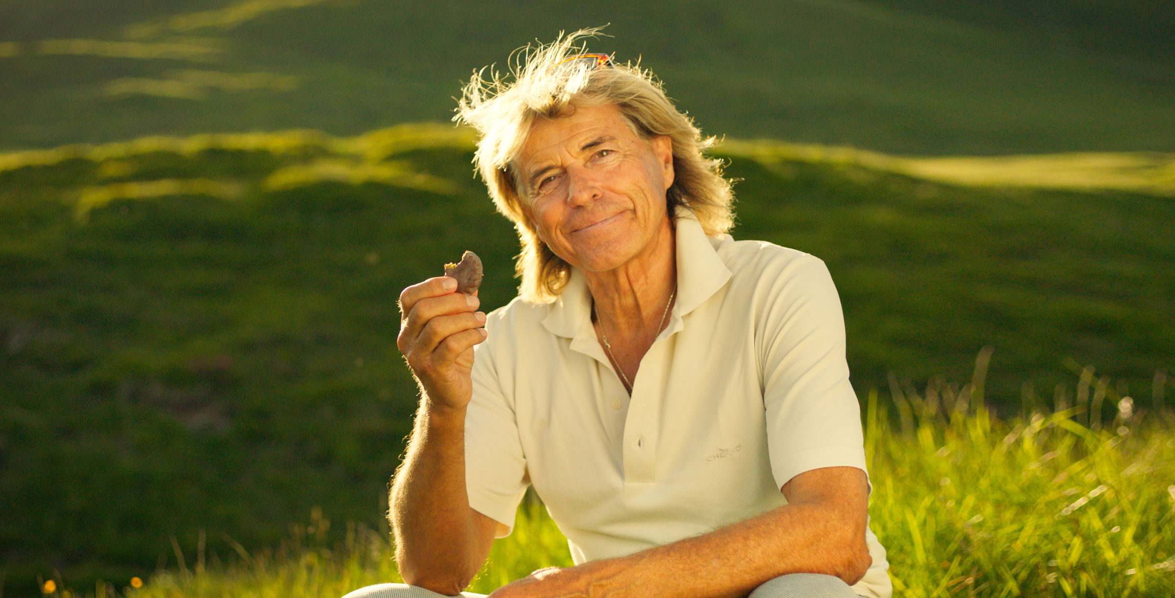 Hansi Hinterseer sitzt in einer grünen Wiese und hält ein SUN Minimeal in der Hand.