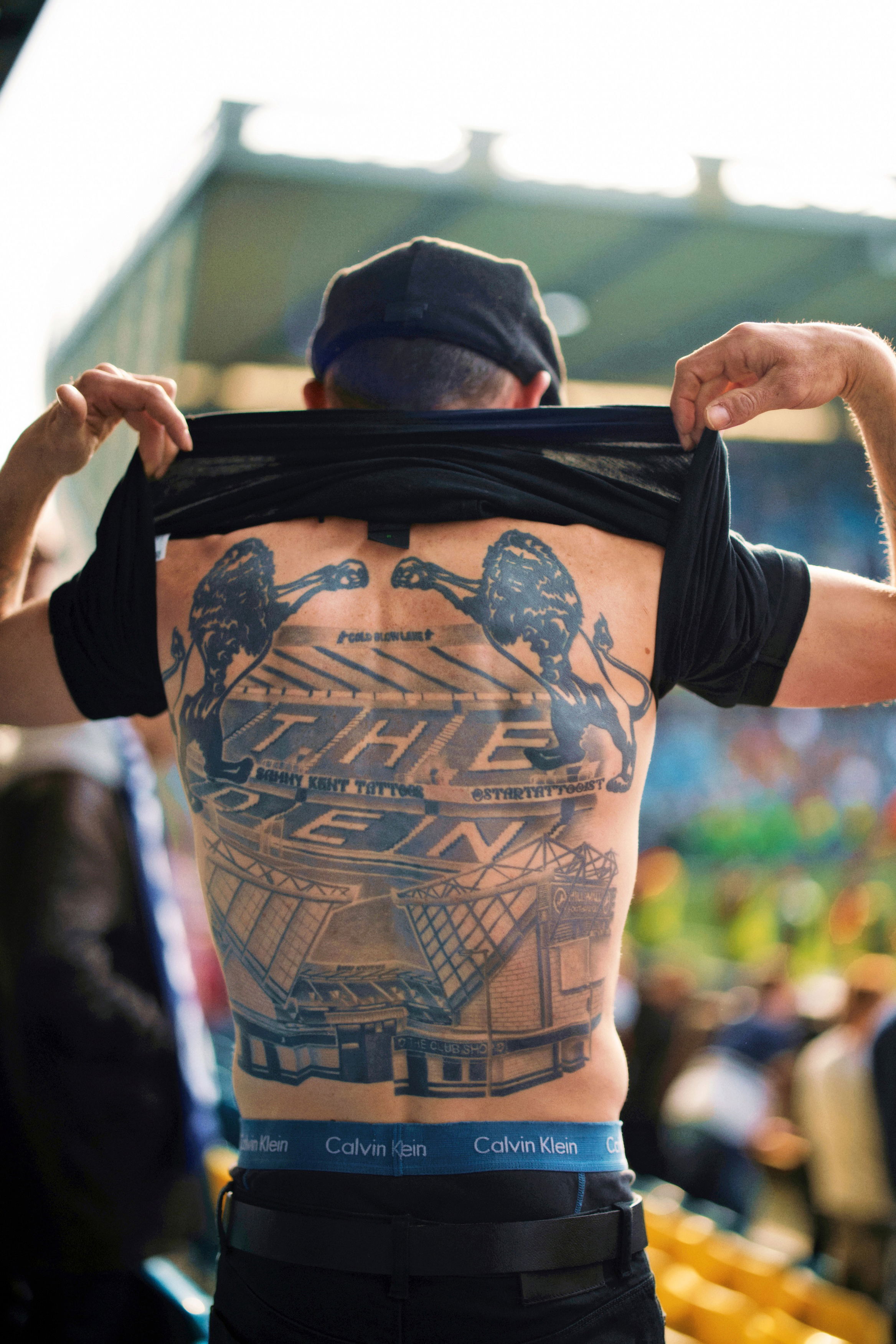 Ein Mann im Stadion hebt sein schwarzes T-Shirt hoch und zeigt ein großflächiges Rücken-Tattoo. Das Motiv zeigt zwei brüllende Löwen, den Schriftzug ‚The Den‘ sowie eine detailreiche Darstellung des Stadions des Millwall FC