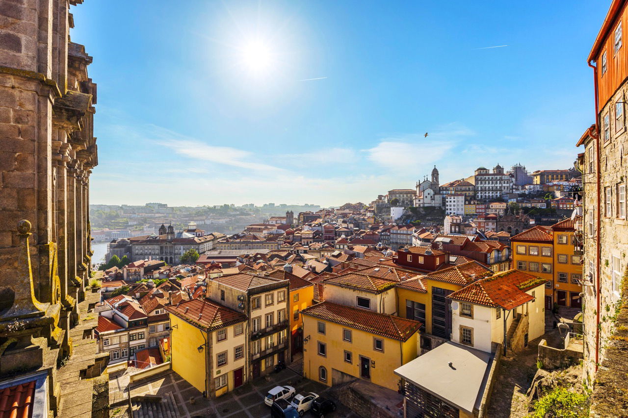 Blick über eine historische Stadt mit dicht aneinander stehenden Häusern mit roten Ziegeldächern, gelben und beigen Fassaden, unter klarem, sonnigem Himmel. Im Hintergrund sind Kirchen und historische Gebäude auf einem Hügel zu sehen