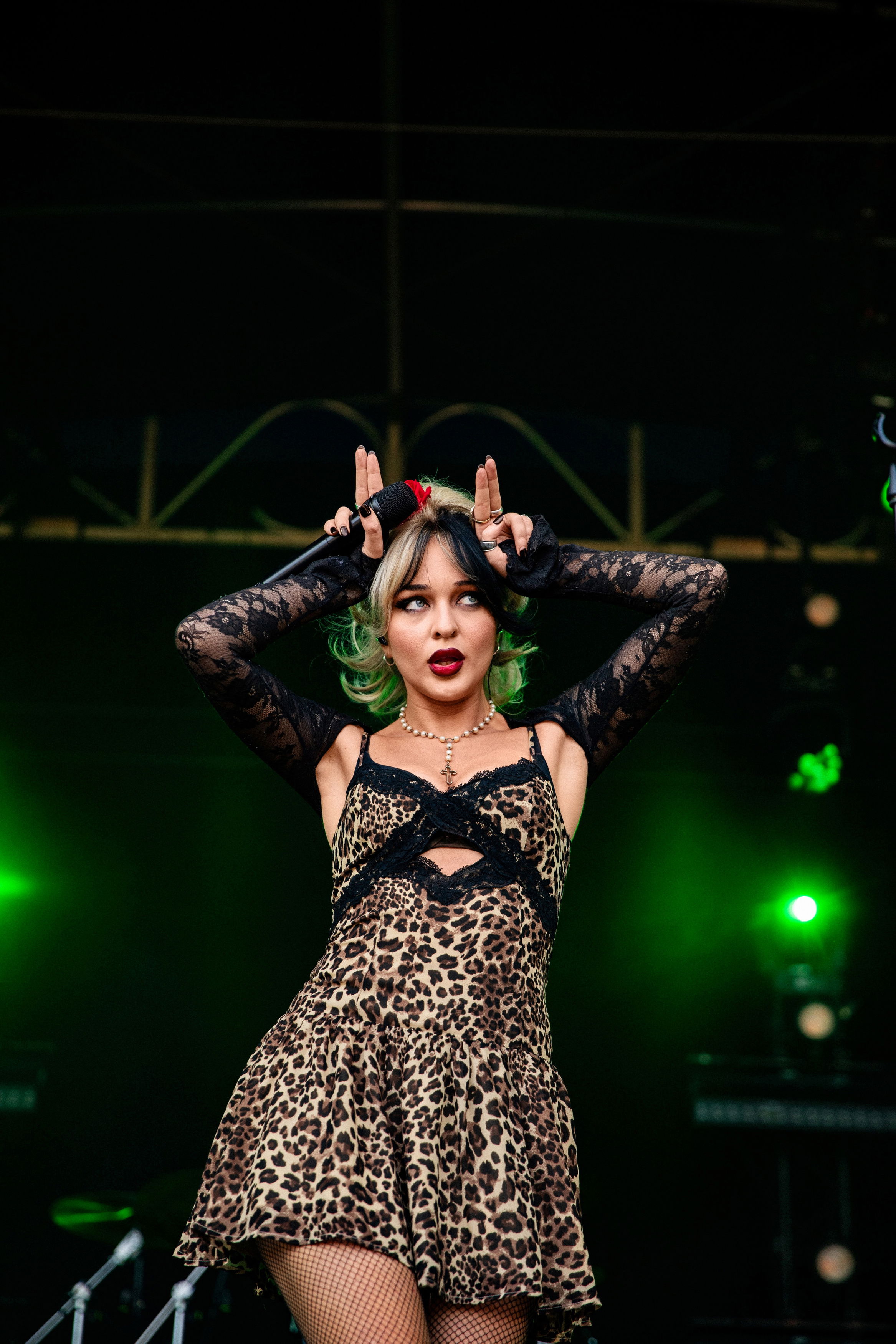 Frau mit zweifarbigem Haar und Leopardenkleid steht auf Bühne, hebt beide Hände mit gespreizten Fingern an den Kopf; grünes Bühnenlicht im Hintergrund.