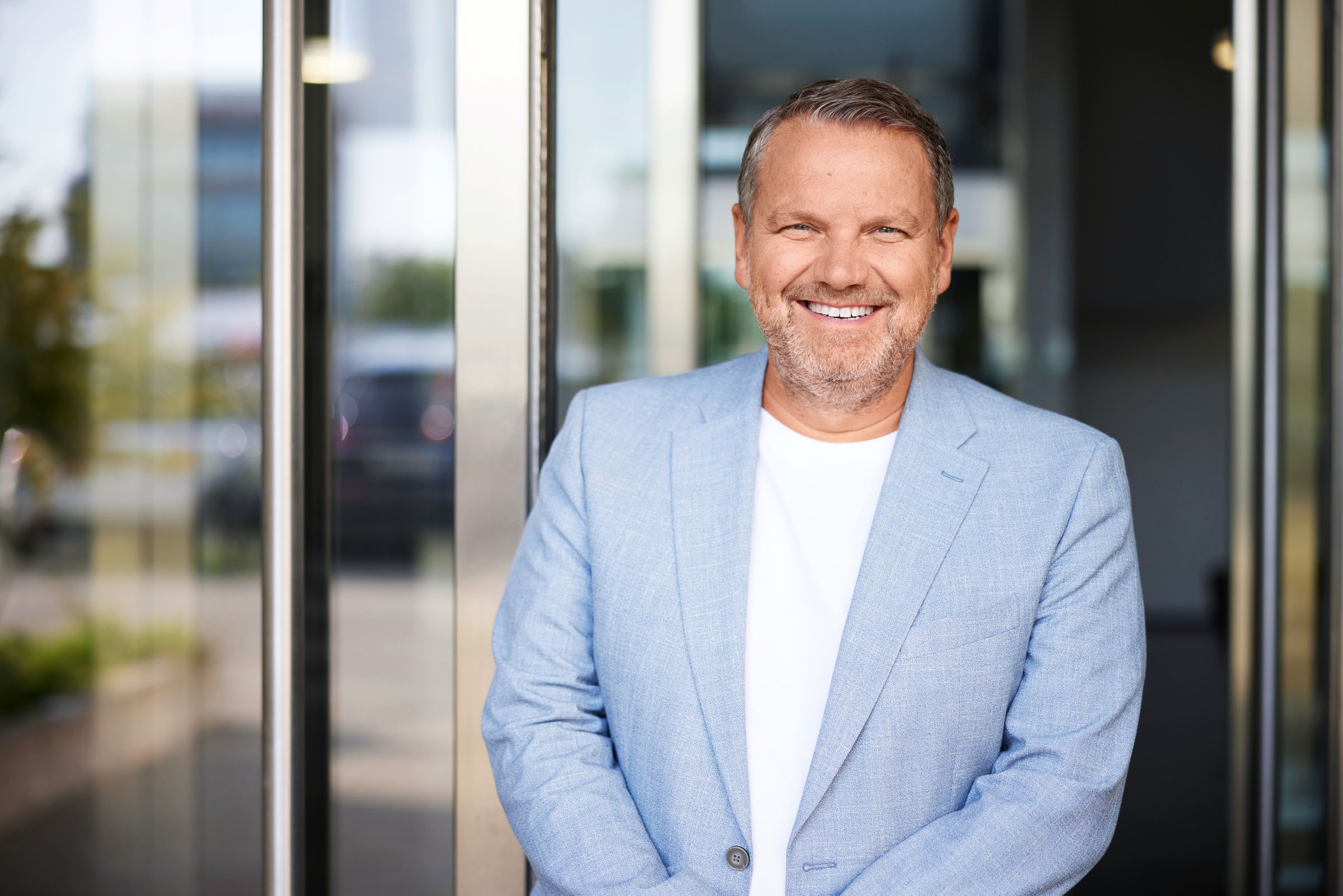 Udo Heuser steht lächelnd steht vor einem Glasgebäude. Er trägt ein hellblaues Sakko über einem weißen T-Shirt und schaut direkt in die Kamera.