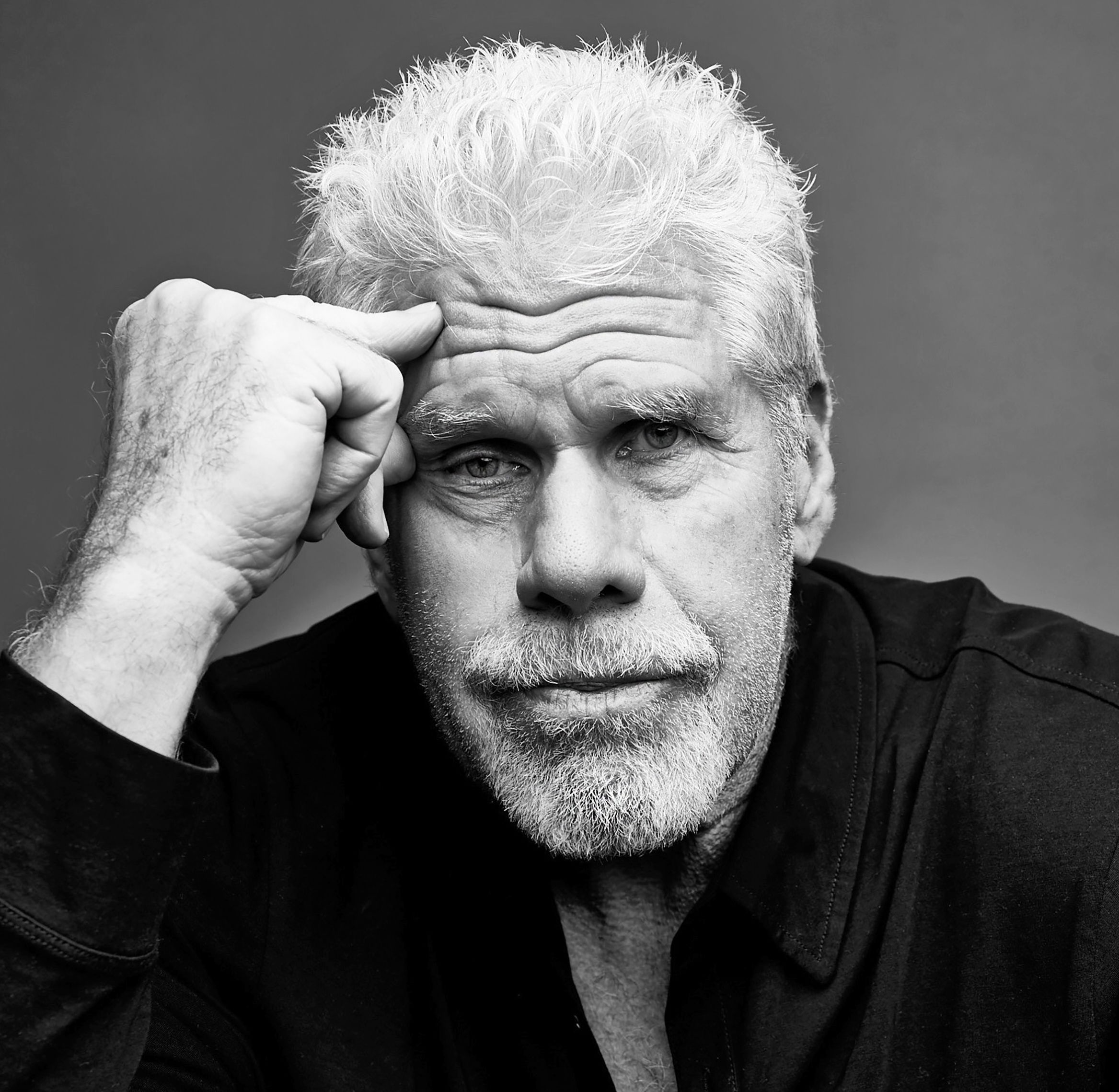 Ein Schwarzweiß-Porträt von Ron Perlman in Nahaufnahme. Er hat dichtes, hellgrau/weißes, leicht zerzaustes Haar und einen kurzen grauen Bart. Der Blick ist direkt in die Kamera, ruhig und ernst, mit leicht zusammengezogenen Brauen. Seine linke Hand liegt an der Stirn/Schläfe, als würde er nachdenken; die rechte Hand ist vorne im Bild zu sehen, mit einem Ring am Finger.