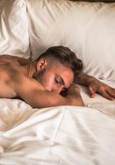 Junger Mann mit nacktem Oberkörper liegt auf dem Bauch schlafend in weißer Bettwäsche