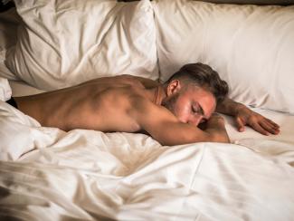 Junger Mann mit nacktem Oberkörper liegt auf dem Bauch schlafend in weißer Bettwäsche