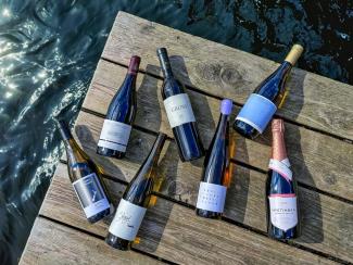 7 einzelne Weinflaschen liegen auf einem Steg über glitzerndem See