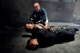 Zwei gefesselte Männer liegen auf Boden in dunklem Raun, daneben kniet entschlossen blickender Mann 