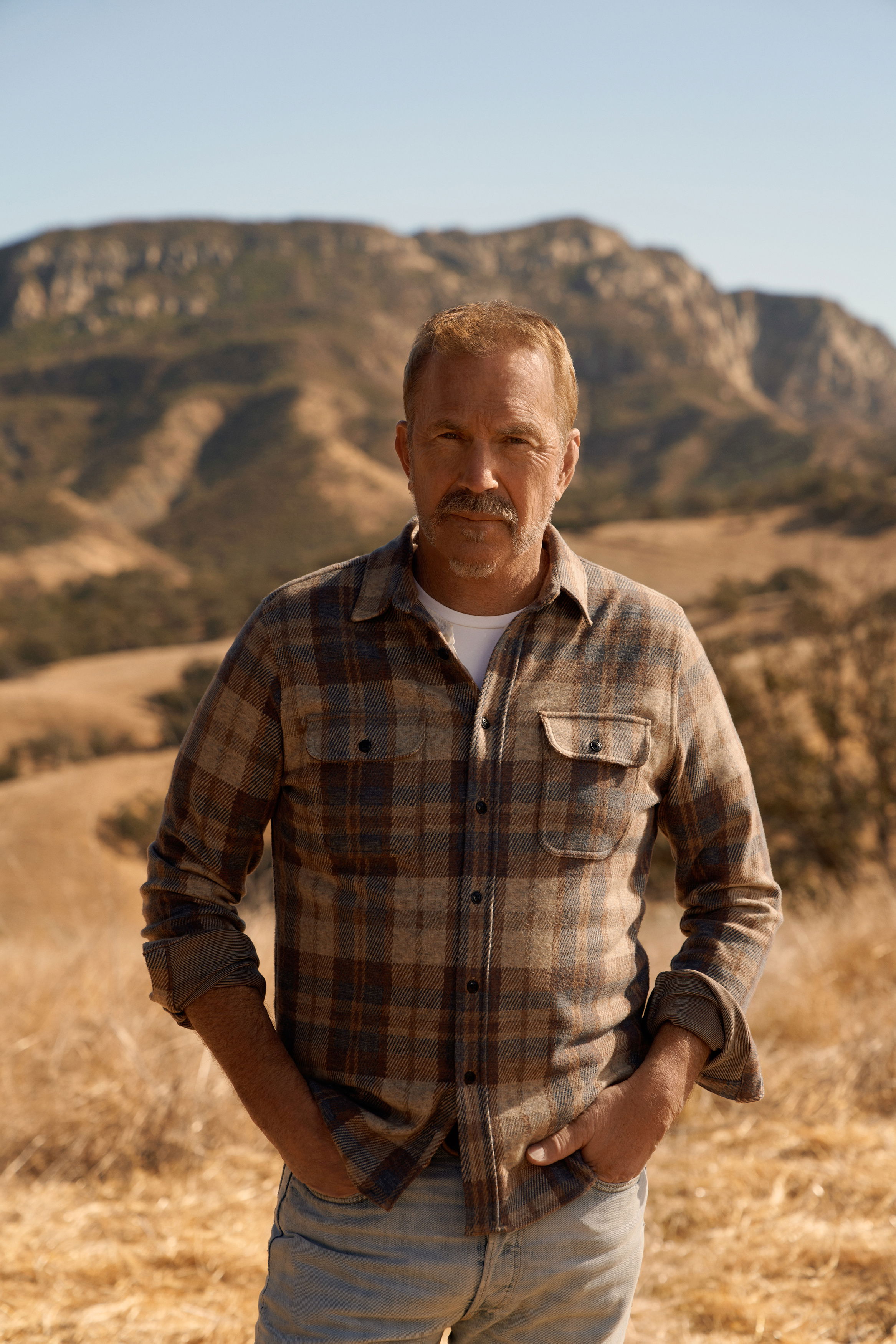 Der Schauspieler Kevin Costner steht in einer kargen Wüstenlandschaft in kariertem Hemd und Jeans